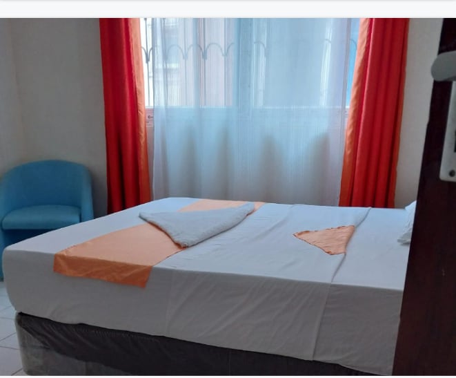 A well-lit sleeping area is presented, featuring a spacious bed adorned with decorative pillows. Bright orange curtains frame a window, allowing natural light to softly illuminate the room. A cozy chair in blue adds a touch of color to the relaxed environment.