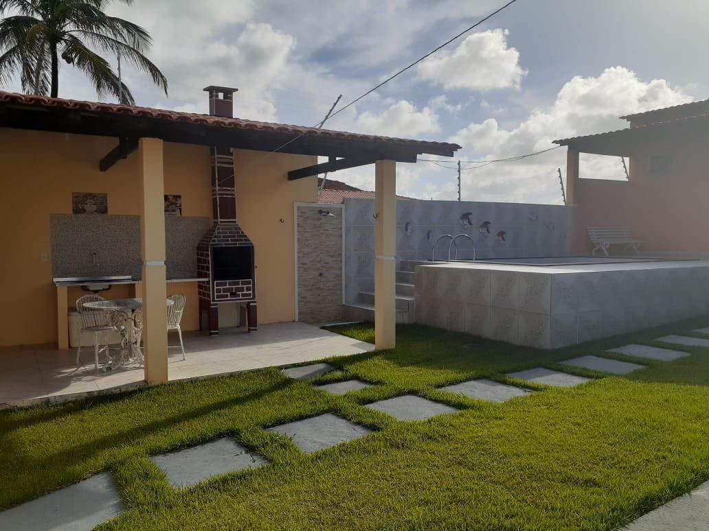 An outdoor area is presented, featuring a barbecue grill and a dining space with two chairs. A rectangular pool is seen adjacent to a stone wall decorated with subtle artwork. Lush green grass and stepping stones create a welcoming pathway, under bright skies.
