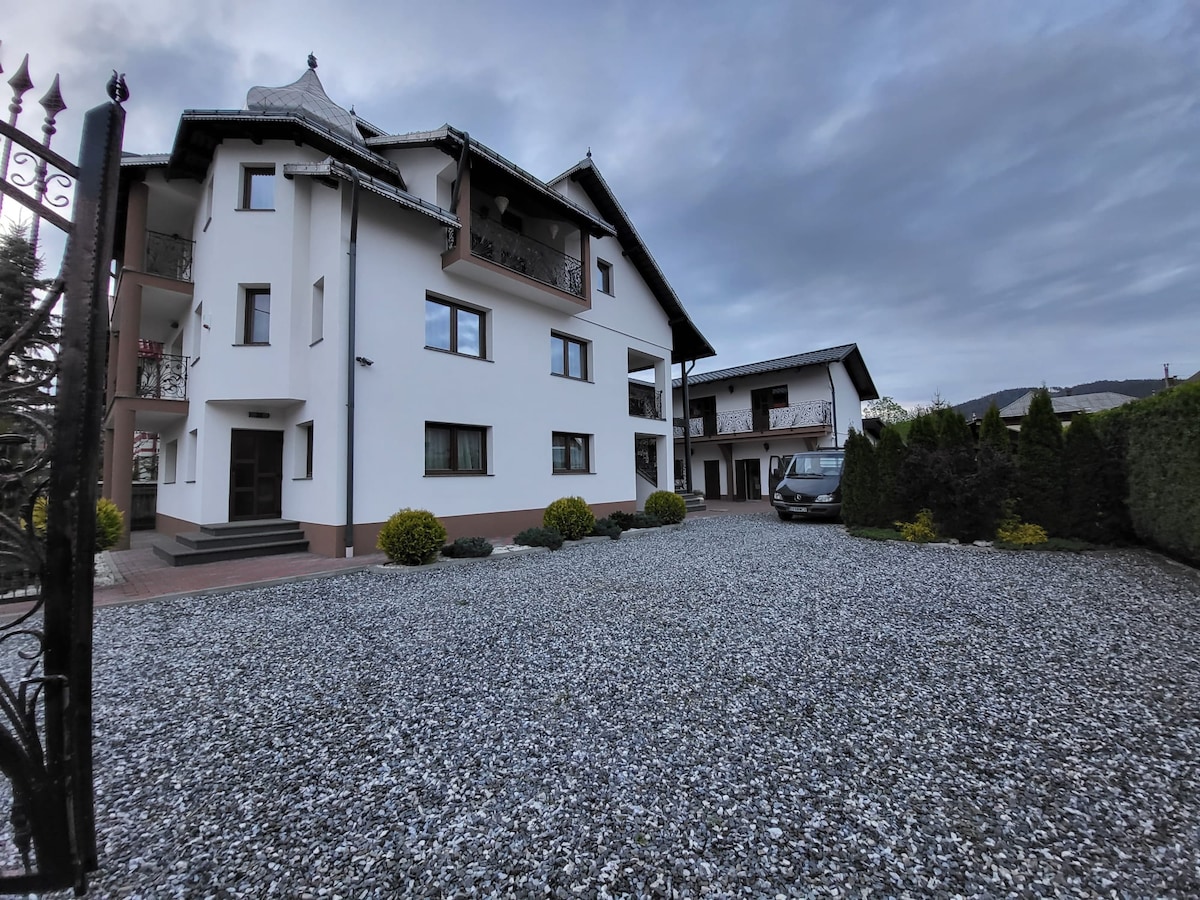 A multi-story building is visible, featuring a mix of traditional and modern architectural elements. The exterior is painted in light colors with dark accents. A gravel driveway leads to the entrance, surrounded by neatly trimmed bushes and small decorative shrubs.