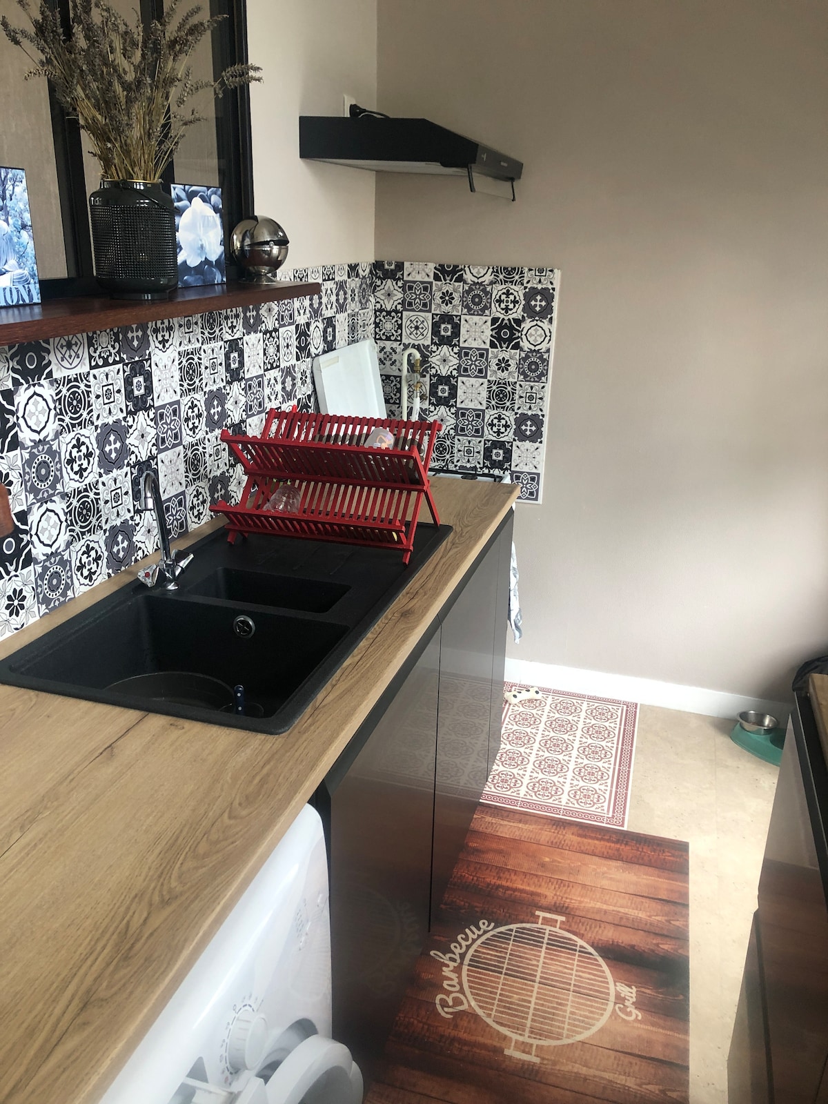 A well-equipped kitchen area is showcased, featuring a sleek black sink and wooden cabinetry. Decorative tiled backsplash adds visual interest. A red dish rack is positioned above the sink, with beige flooring and a patterned rug. The space appears organized and functional.