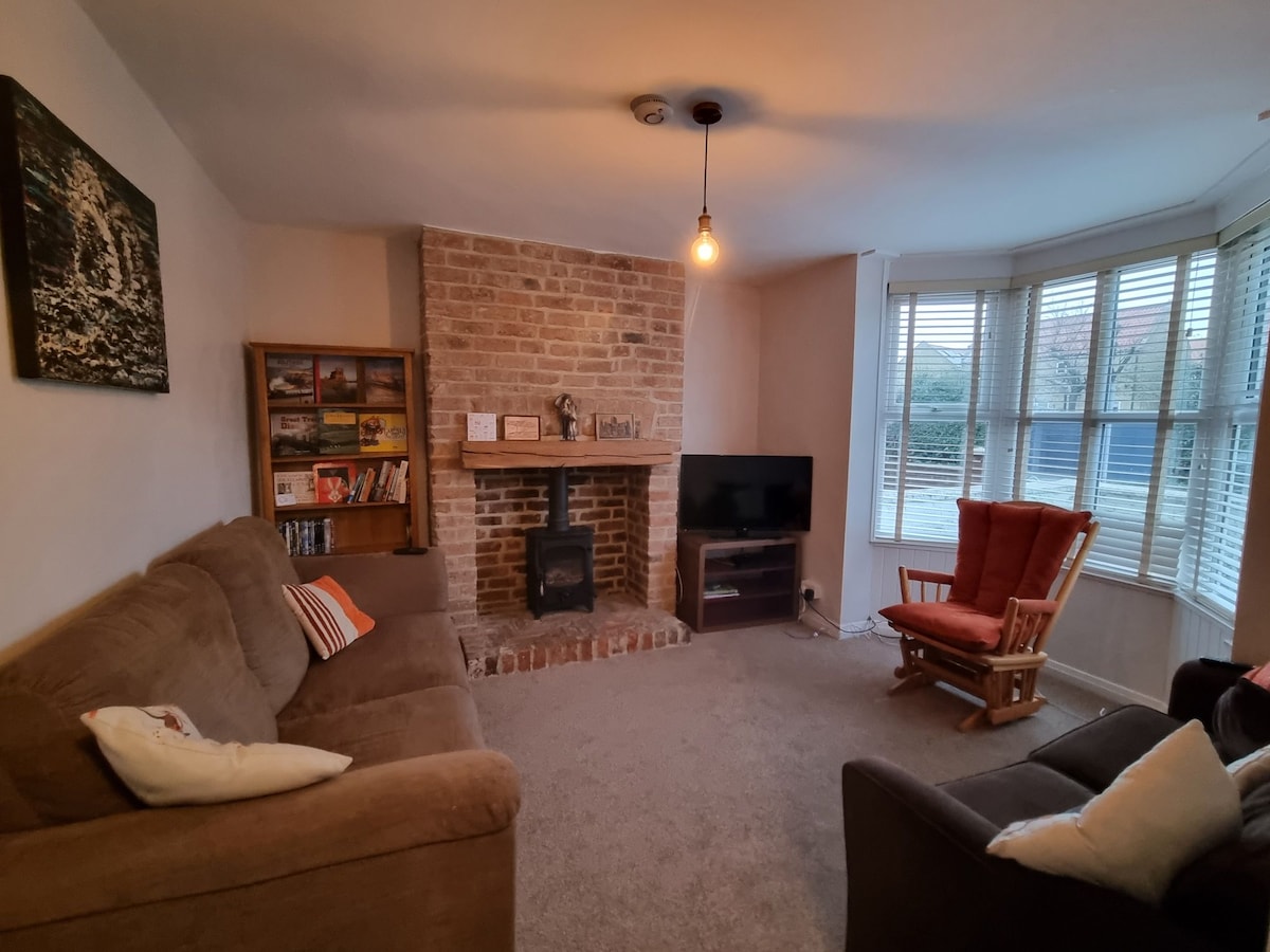 A cozy living room features a stone fireplace with a wood burning stove and a flat-screen TV. Comfortable seating includes a brown sofa, a rocking chair, and an armchair, arranged to encourage conversation. Natural light pours in through large windows adorned with wooden blinds.