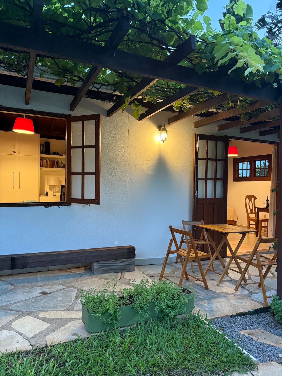 The exterior features a charming patio area with folding chairs and a small table, surrounded by greenery. A vine-covered trellis provides shade, while warm lighting enhances the inviting entrance to the interior space, visible through the open door.