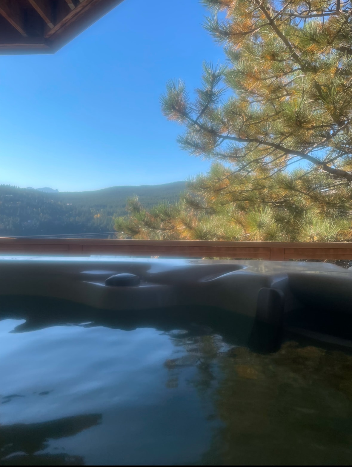 A hot tub is positioned on a balcony, surrounded by tall pine trees. The clear blue sky and distant mountains create a serene backdrop, reflecting in the still water. The area offers an inviting space for relaxation, with an emphasis on nature's beauty.