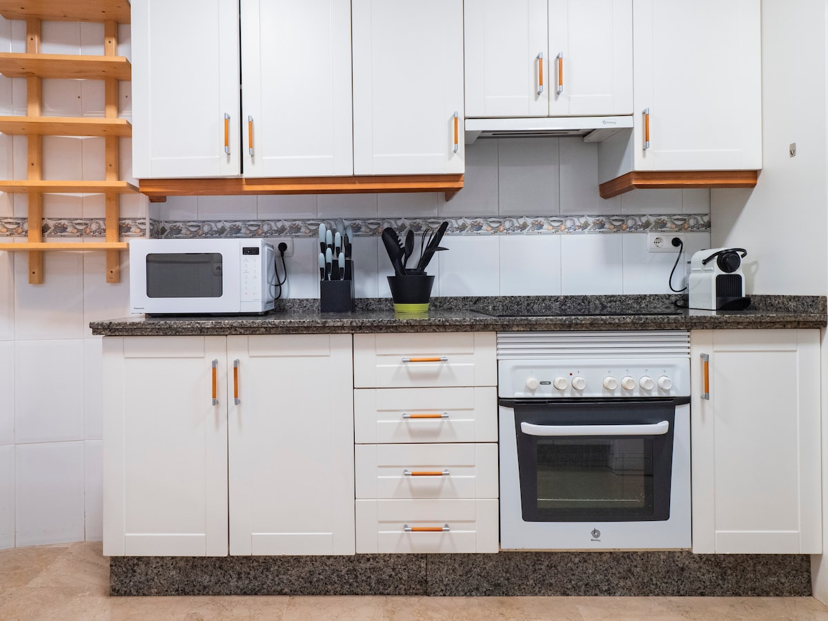A modern kitchen is equipped with essential appliances, including a microwave, coffee maker, and oven. Cabinets with sleek white doors are complemented by a dark granite countertop. A set of utensils is neatly organized in a holder, while a wooden shelf provides additional storage.