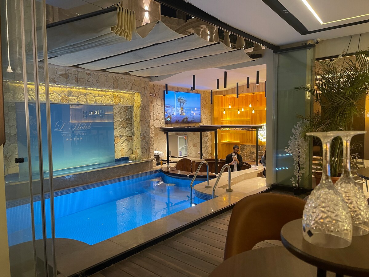 An indoor swimming pool is framed by glass walls, providing a clear view of the water. Lounge areas are set alongside the pool, with modern furniture and soft lighting enhancing the ambiance. A large screen is mounted on the wall, visible in the background.