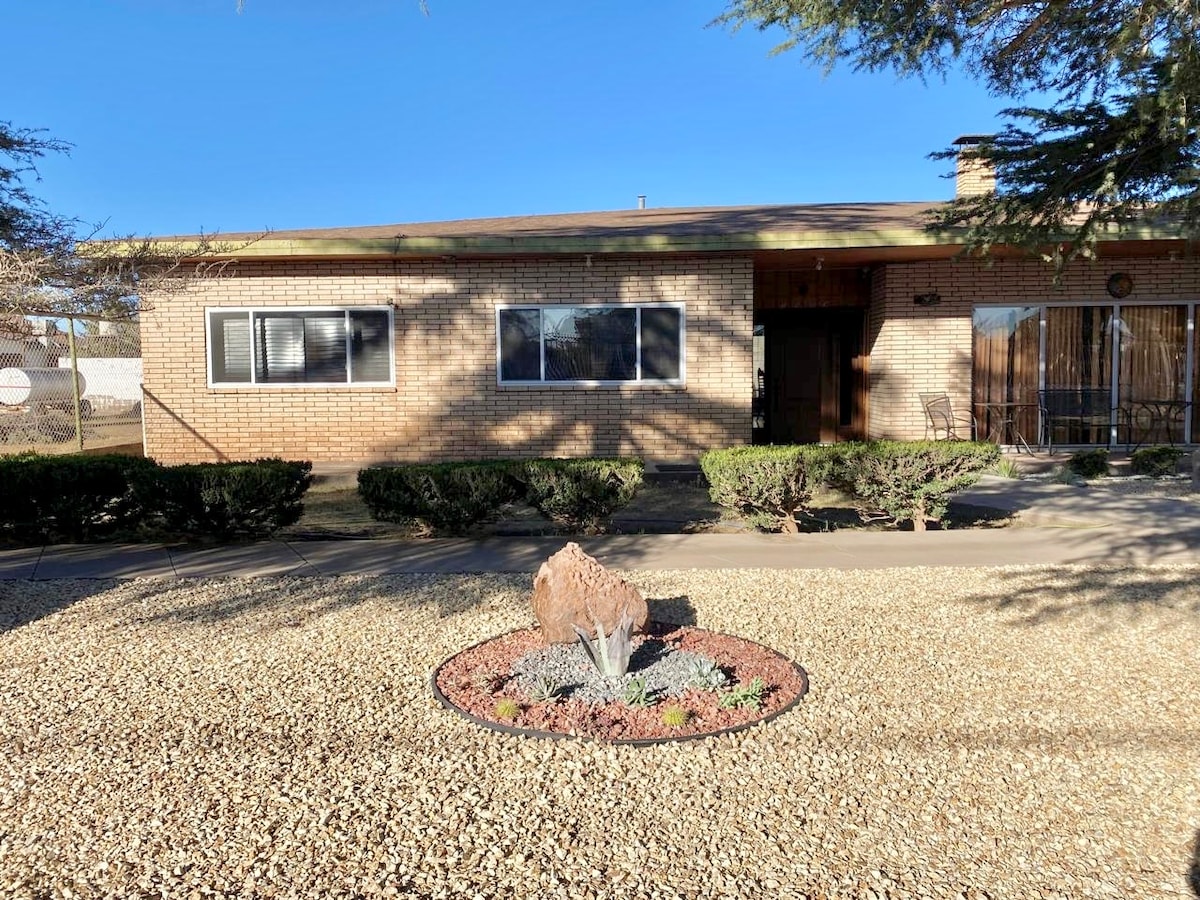 A single-story brick house is presented, featuring large front windows and a welcoming entrance. The well-maintained front yard is outlined with neatly trimmed shrubs and a decorative rock garden, providing an inviting space that is well-lit by natural sunlight.