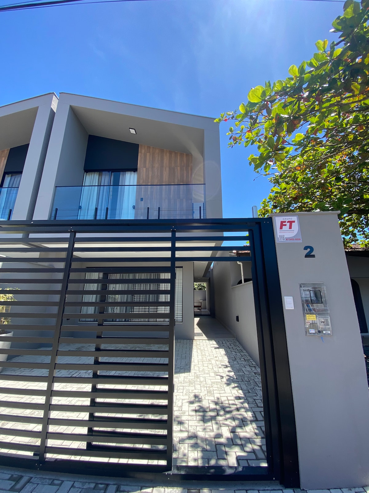 A modern, multi-story building is depicted, featuring a combination of light grey and natural wood accents. The entrance is framed by a black horizontal gate, leading to a paved pathway. Bright sunlight highlights the clear blue sky above and the surrounding foliage.