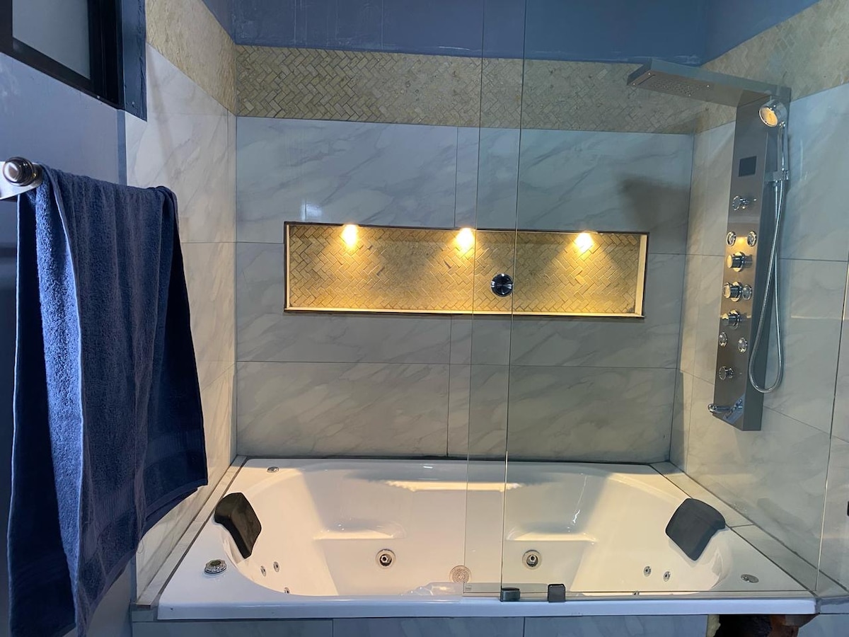 A modern bathroom features a spacious jacuzzi tub with built-in jets, enclosed by a glass partition. Soft lighting is provided by three small wall fixtures, illuminating the elegant tile work surrounding the tub. A towel is neatly hung on a nearby rack.