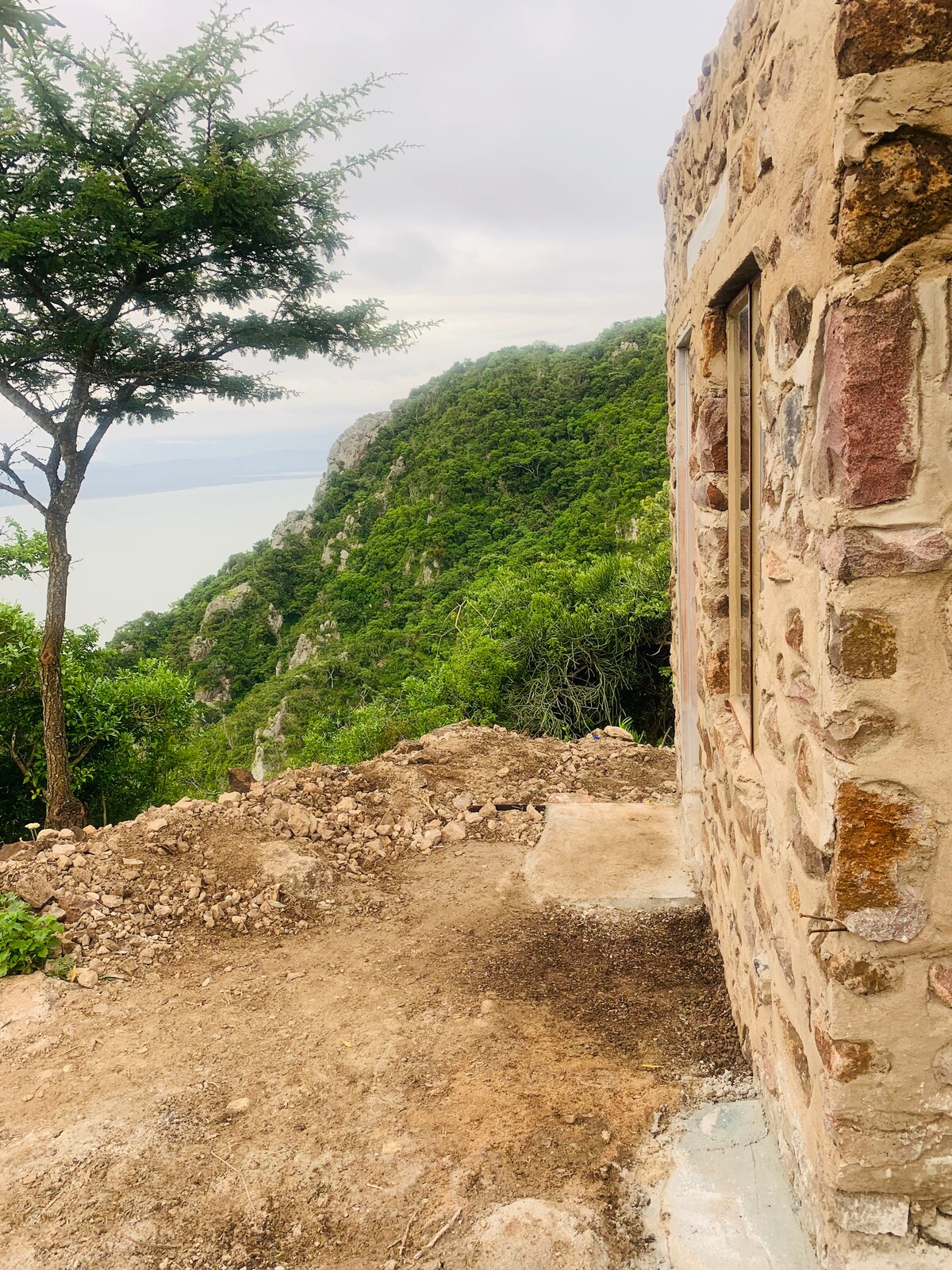 A rustic stone structure is positioned on a hillside, offering views of a lush green landscape and a body of water in the distance. The surrounding earth is uneven, with patches of vegetation and rocky terrain leading to a natural edge.