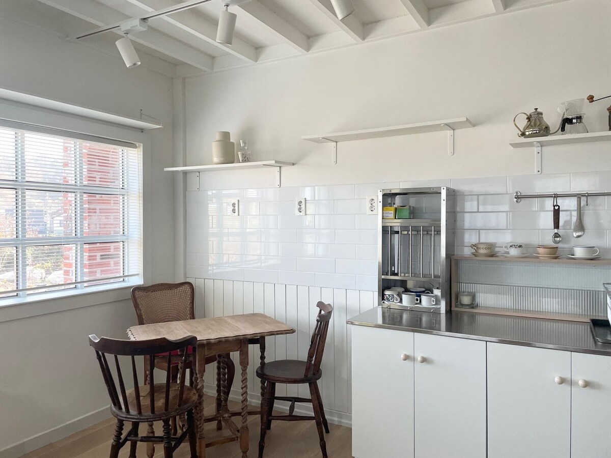 A small kitchen area features a wooden table accompanied by two chairs, one with a woven back. White tiled walls create a clean backdrop, complemented by open shelves displaying kitchenware. Natural light enters through a window, enhancing the bright and airy atmosphere.