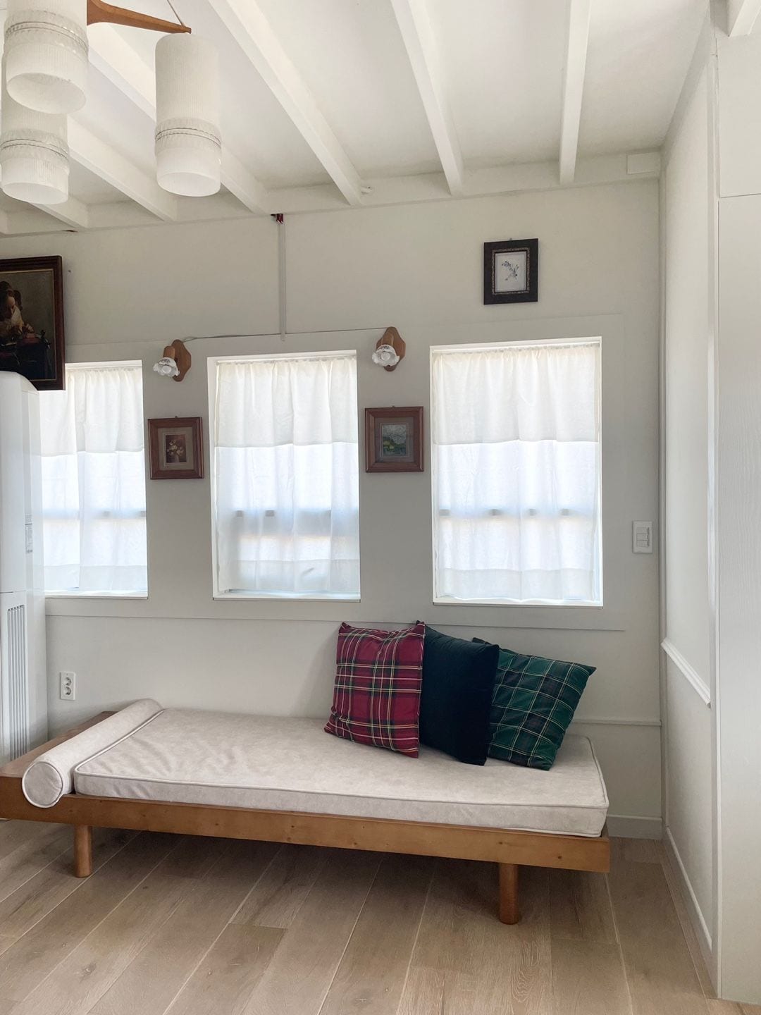 A cozy nook features a wooden daybed with a light cushion, adorned with colorful plaid pillows. Natural light softly filters through three sheer-curtained windows, illuminating the walls decorated with framed art pieces and adding a sense of warmth to the space.