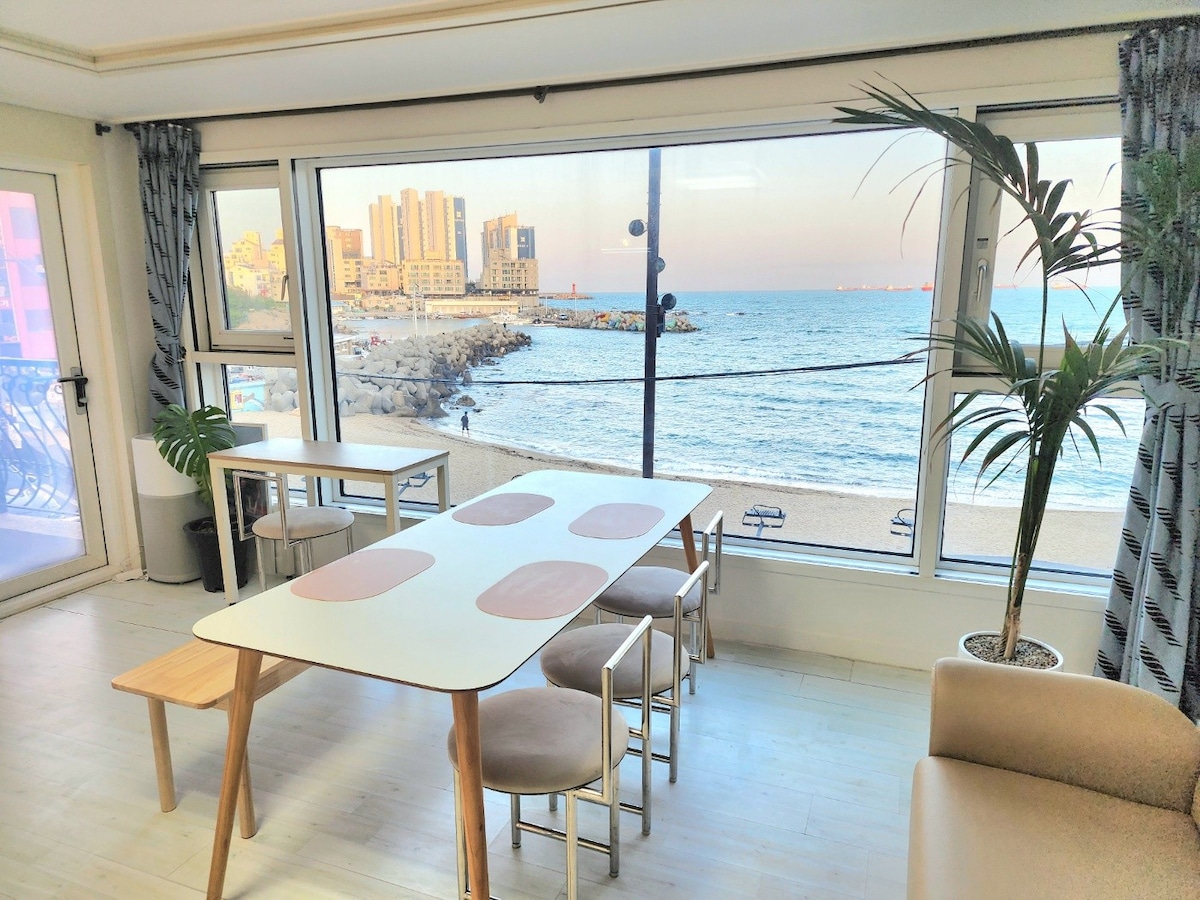 A dining area features a light-colored table surrounded by four chairs, complemented by a potted plant. Large windows overlook the beach and ocean, offering views of the shoreline and distant buildings, creating a bright and airy atmosphere.