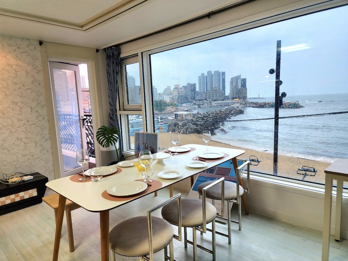 A dining area is shown with a modern table set for six. Large windows overlook the beach and ocean, allowing for plenty of natural light. The distant city skyline can be seen through the glass, and soft, neutral tones create a calm setting.