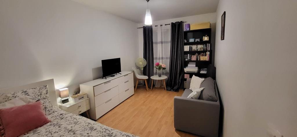 A well-lit bedroom features a spacious bed adorned with floral bedding. A television sits on a dresser, while a small round table is paired with two chairs. A bookshelf displays a variety of books, and large curtains frame the window, allowing natural light to filter through.