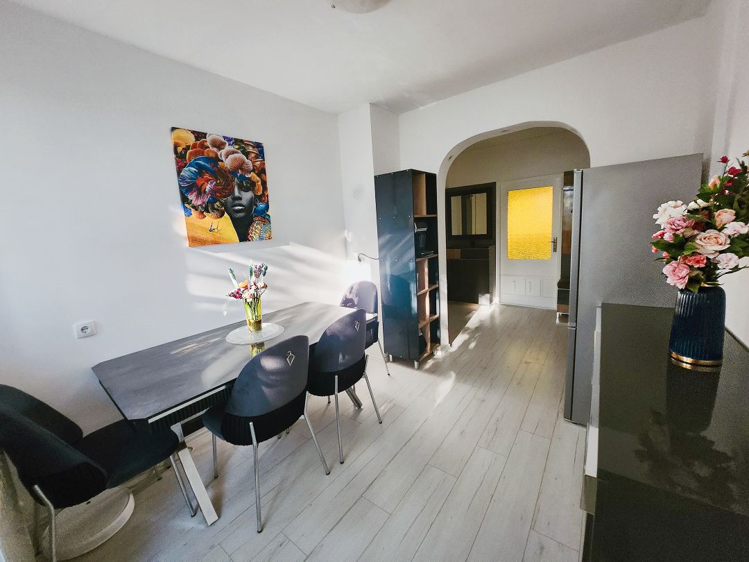 A dining area features a black table surrounded by six chairs. Natural light enters through an arched doorway, highlighting a colorful wall art piece. A dark cabinet stands beside a bookshelf, both contributing to the room's modern design.