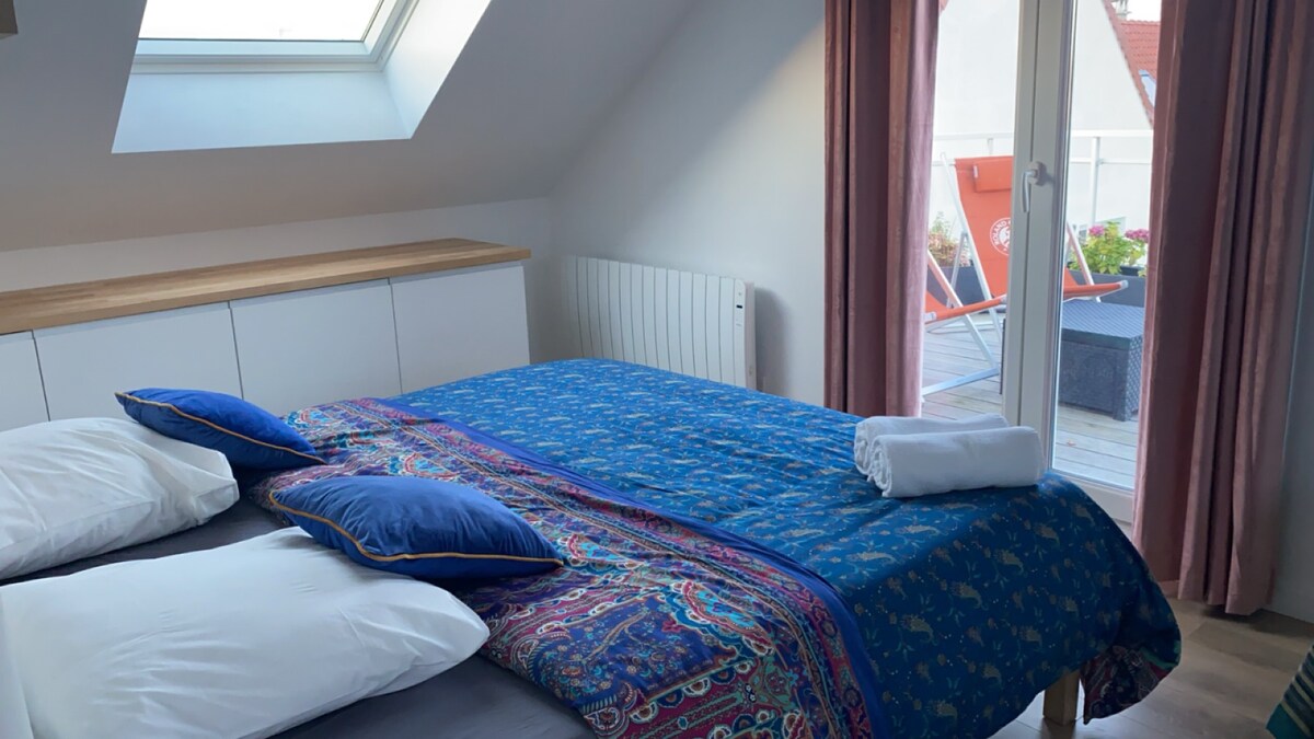 A bright bedroom is featured, furnished with a spacious bed draped in a patterned blanket. Two pillows lie atop the bed, and a skylight allows natural light to fill the room. A door leads to a terrace with outdoor seating.