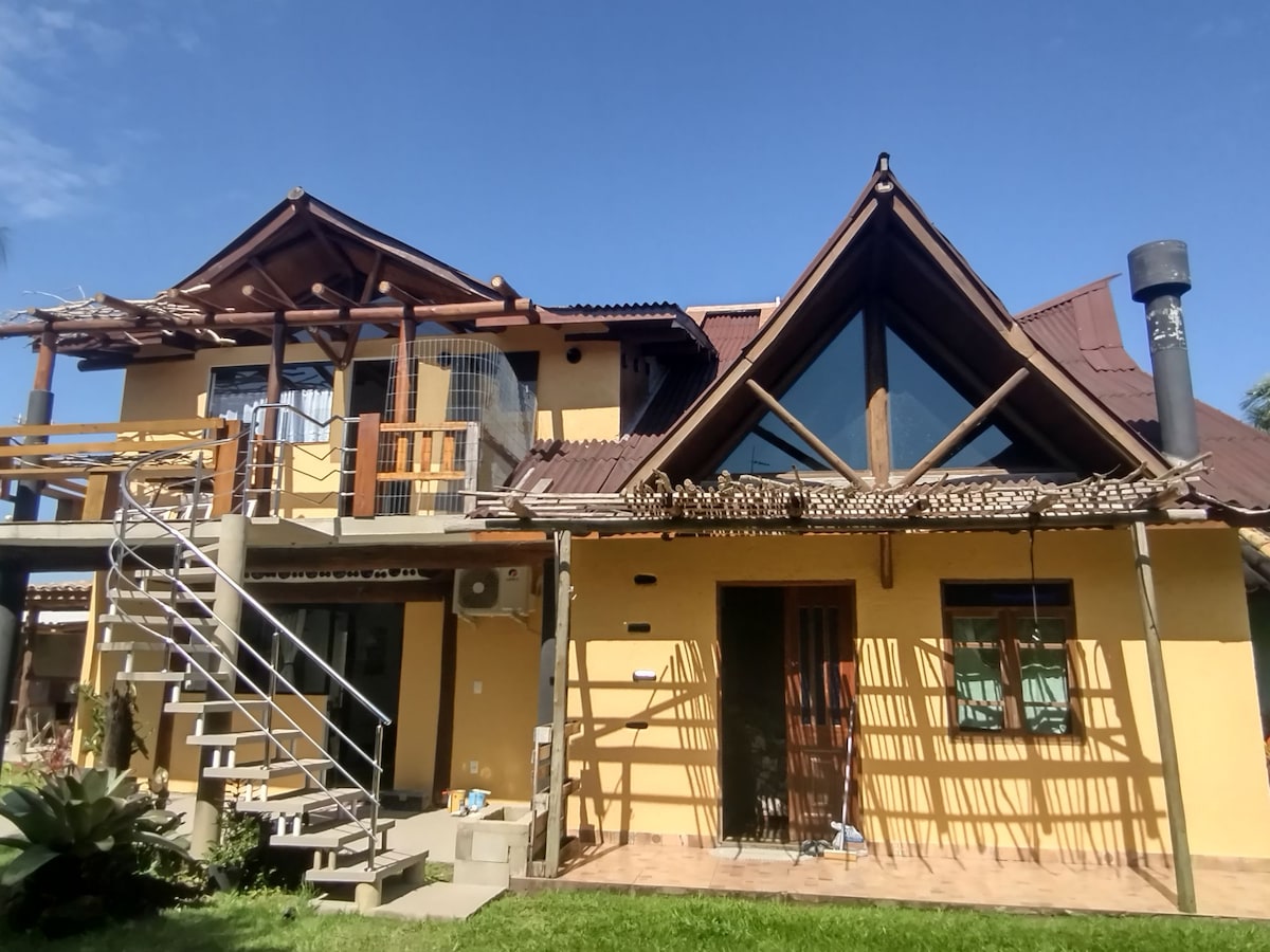 The exterior of a two-story home displays a blend of natural materials and modern design elements. A prominent triangular roof and large windows enhance natural light. A staircase leads to a balcony, while textured walls reflect a unique architectural style in a well-maintained garden setting.