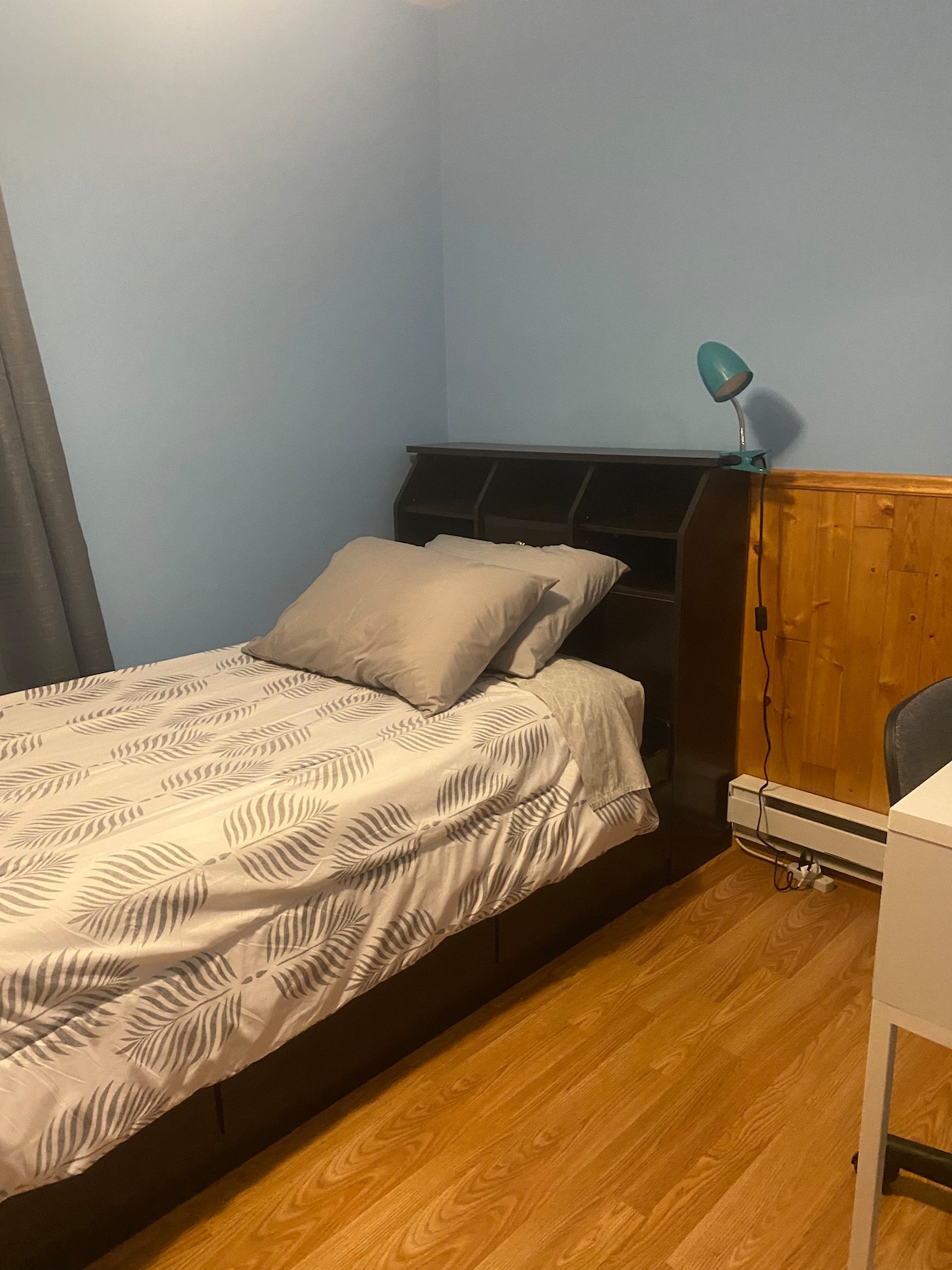 A small room is presented, featuring a captain's twin bed with a light-patterned blanket and two pillows. A wooden shelf is attached to the wall above the bed. A desk and chair are positioned near the bed, complemented by a lamp on the shelf. Grey curtains adorn the window.