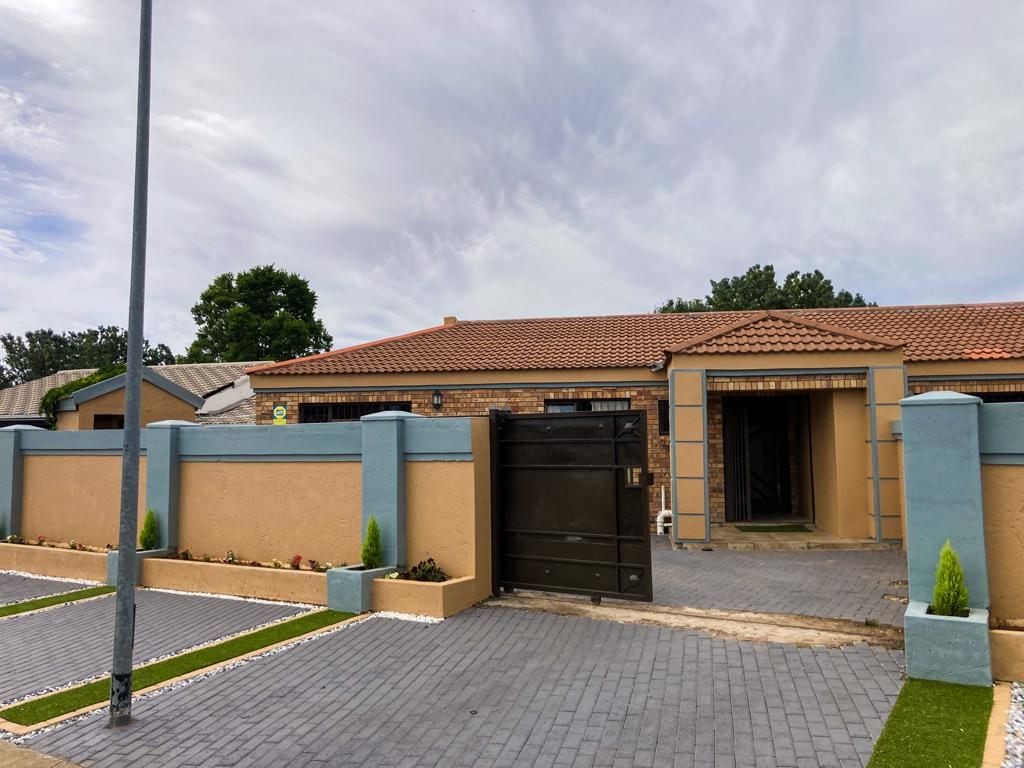 The exterior of the property features a modern facade with a combination of light brown walls and blue accents. A large black gate provides entrance, while a well-maintained driveway lined with green grass leads up to the house. Surrounding vegetation adds to the inviting appearance.