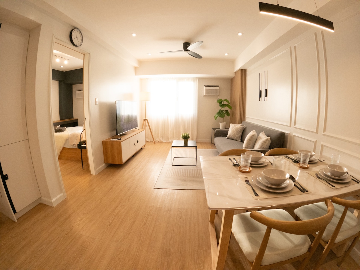 This open living area features a comfortable sofa and a coffee table, complemented by a dining table set for four. Natural light flows through the large window, creating a bright ambiance. A sleek television is mounted on the wall, with an indoor plant adding a touch of greenery.