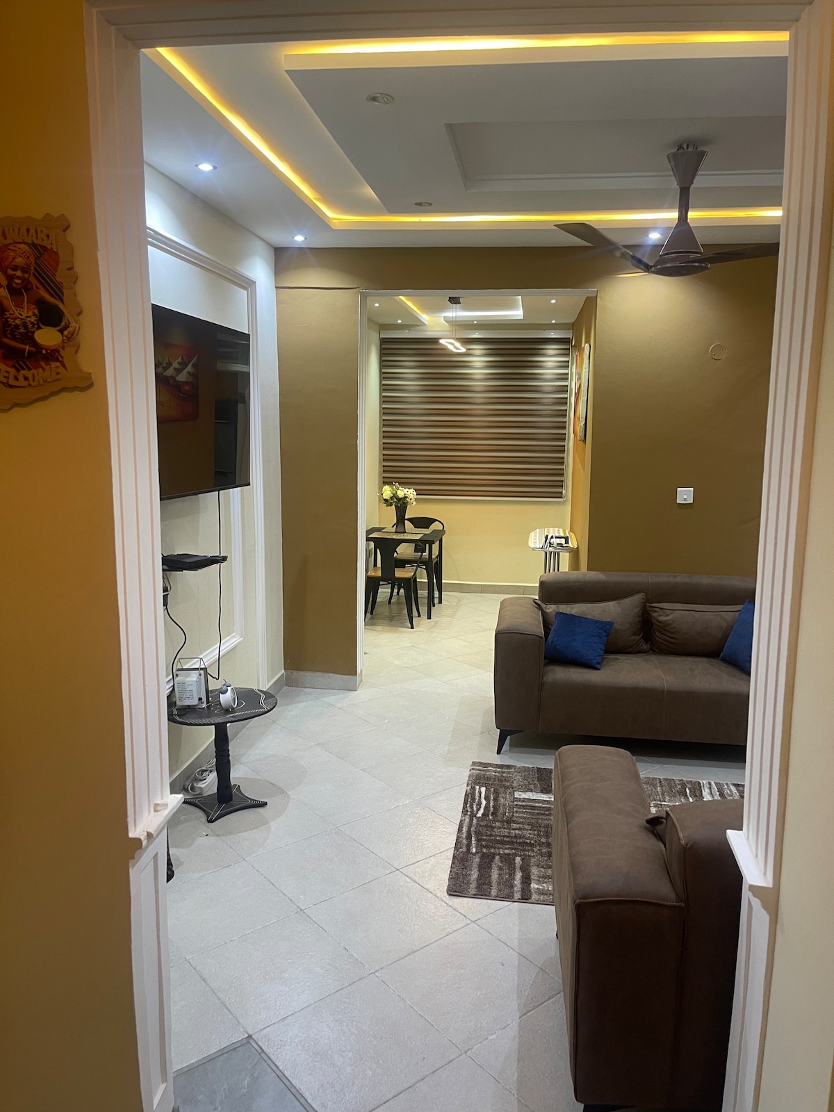 A welcoming entryway leads into a modern living space featuring a cozy sofa set and a small dining area. The room is illuminated by soft ceiling lights, while a decorative fan enhances comfort. A decorative piece hangs on the wall, adding character to the area.