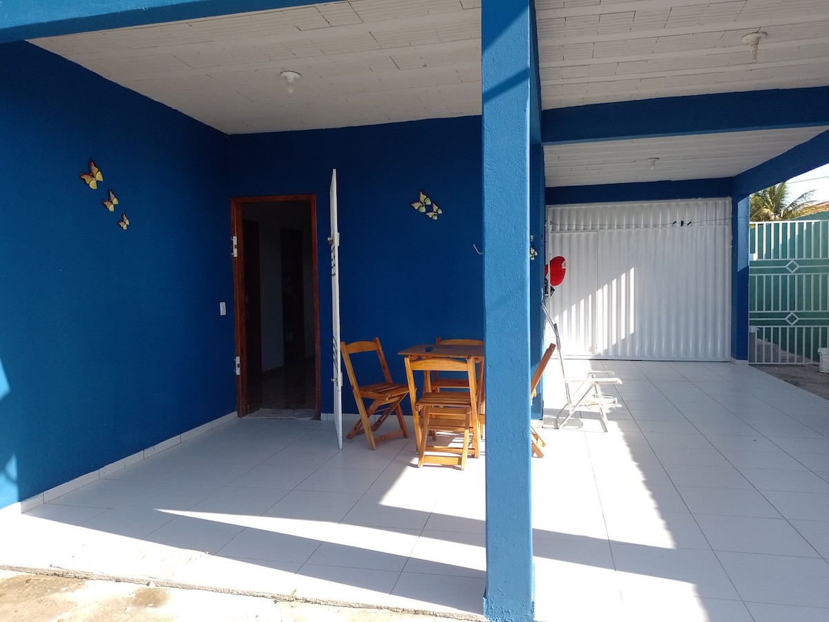 An outdoor patio area features a blue accent wall, complemented by wooden folding chairs and a small table, providing a comfortable space for gatherings. Natural light streams in, highlighting the tiled flooring and the open access to the surrounding area.