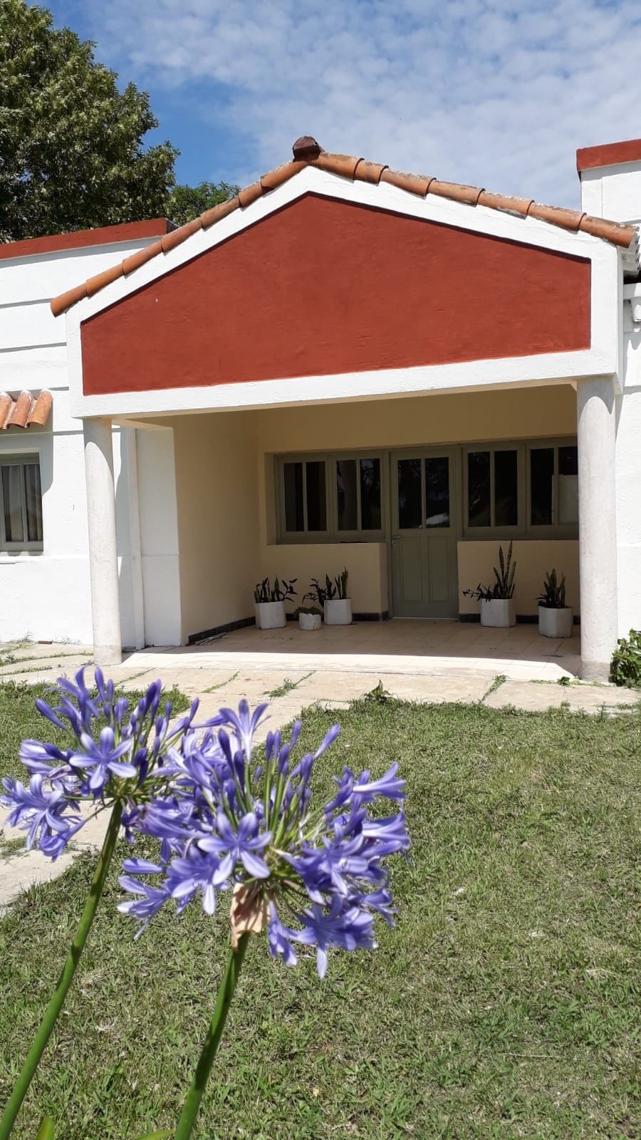 The entrance features a prominently styled gabled roof and is framed by stone columns. Large windows allow natural light to enter, while potted plants add greenery along the pathway leading to the entrance, complemented by purple flowers in the foreground.