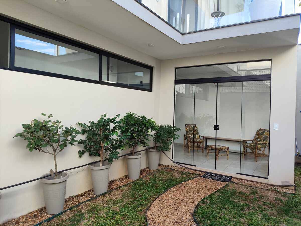 A serene outdoor space features a pathway of pebbles leading to a glass-enclosed area with seating. Surrounding plants in light-colored pots add greenery, while large windows allow natural light to flow in.