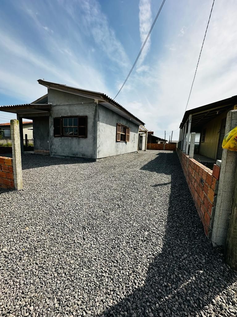 A spacious outdoor area features a gravel surface bordered by low brick walls, leading to a single-story house with a simple facade. Sunlight casts shadows, highlighting the structure's clean lines and multiple windows, creating an open and inviting atmosphere.