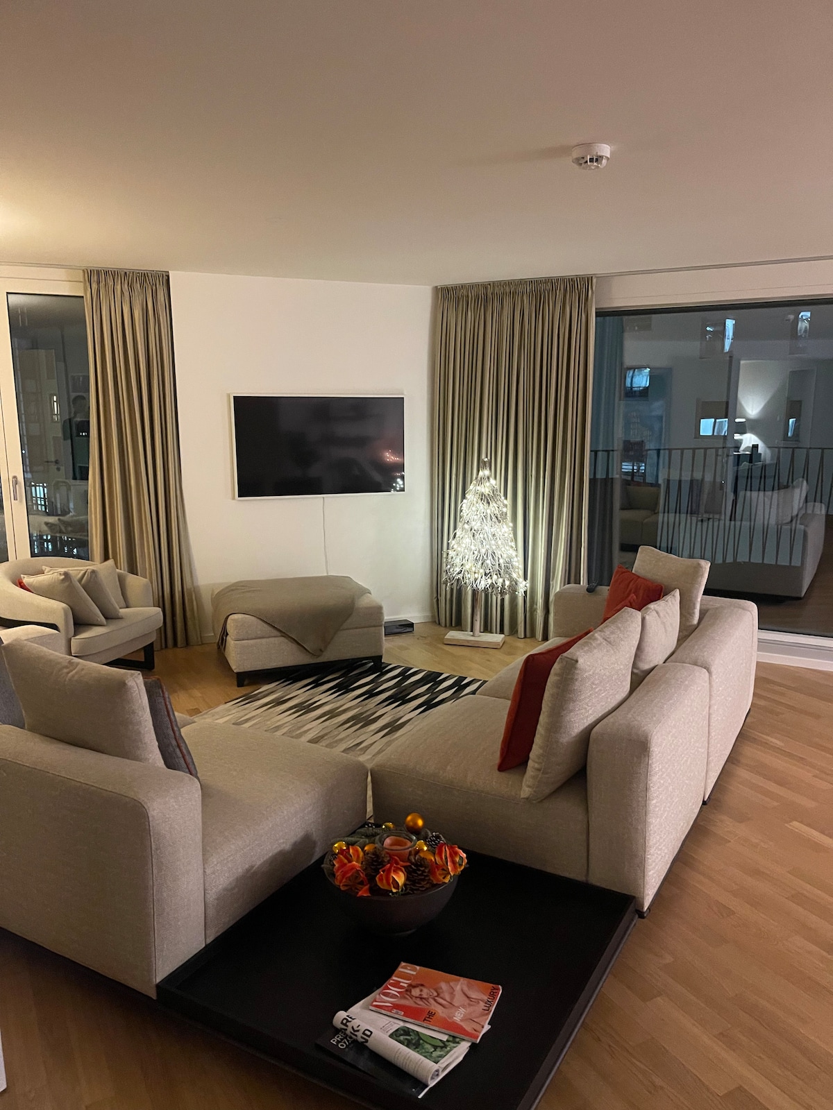 A modern living area is presented, featuring two armchairs and a spacious sofa adorned with decorative cushions. A low coffee table is placed at the center, holding a bowl of flowers. A television is mounted on the wall, with tall curtains framing the large windows.