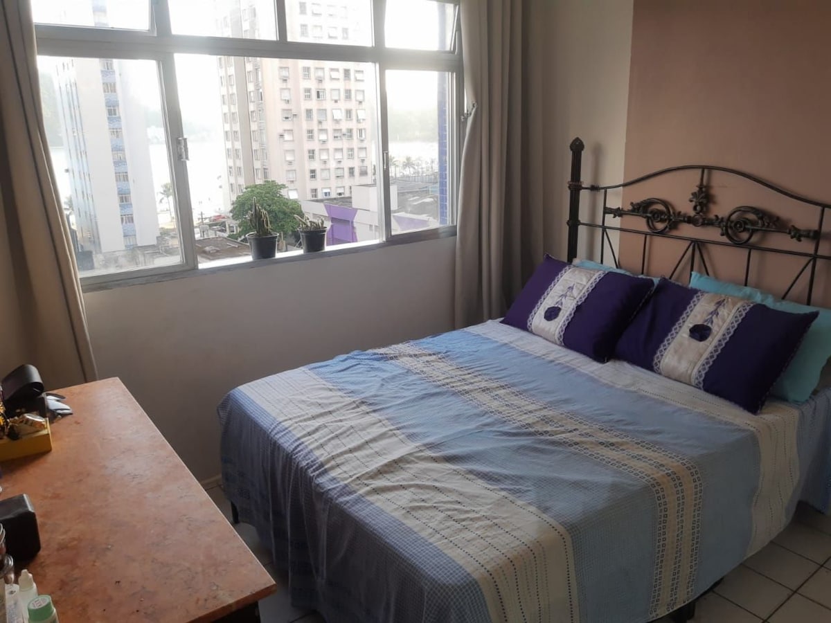 A spacious sleeping area features a comfortable bed with purple-accented pillows, flanked by large windows that offer views of the surrounding cityscape. A wooden desk stands nearby, providing a practical workspace. Natural light illuminates the room, enhancing the serene atmosphere.