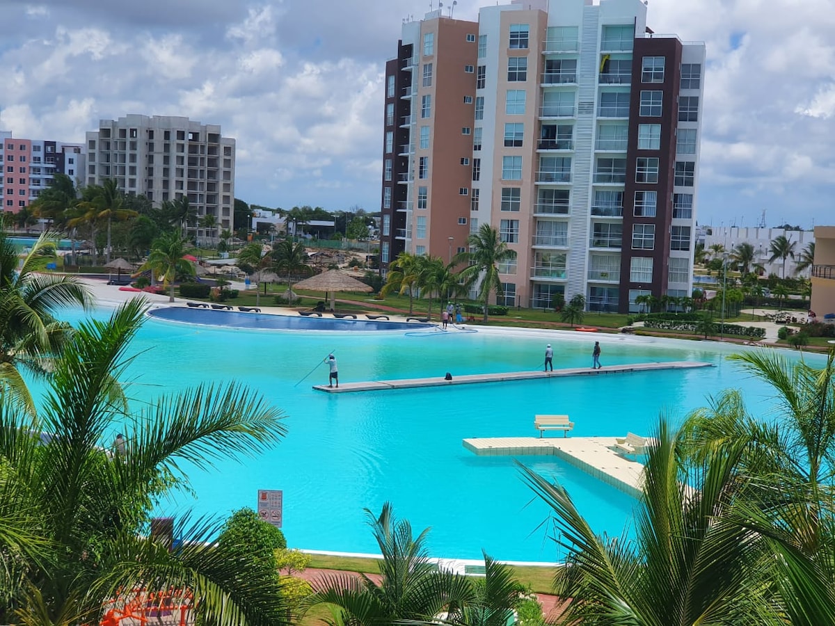 A panoramic view of a large lagoon features calm turquoise water, surrounded by lush greenery. A walking path extends across the lagoon, with individuals enjoying leisure activities. Modern residential buildings rise in the background, contributing to the tranquil atmosphere of the area.