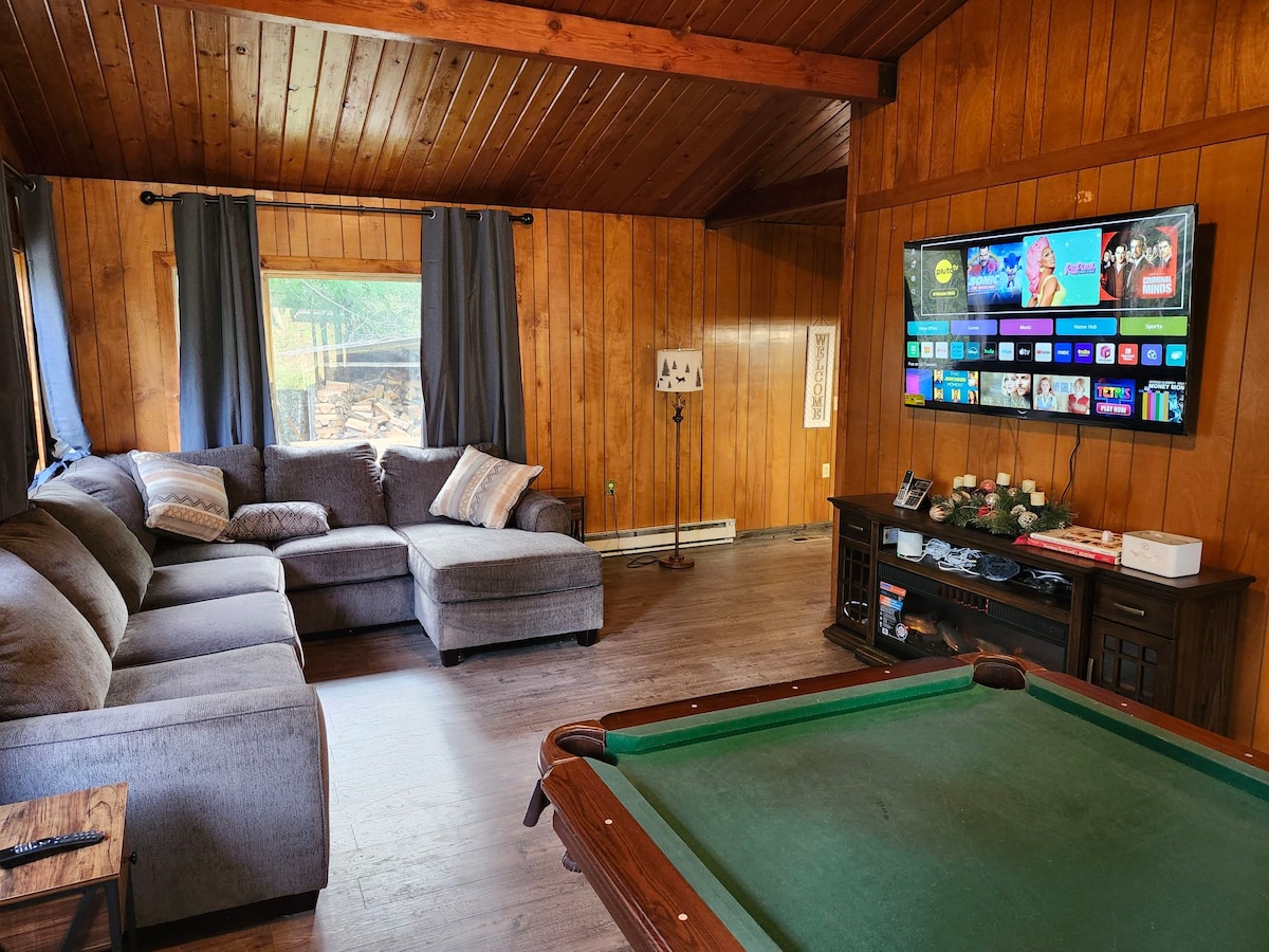 A comfortable living area features a large sectional sofa and a modern television mounted on a wooden wall. Light enters through a nearby window, illuminating the warm wooden paneling. A pool table occupies one corner, adding to the recreational space.