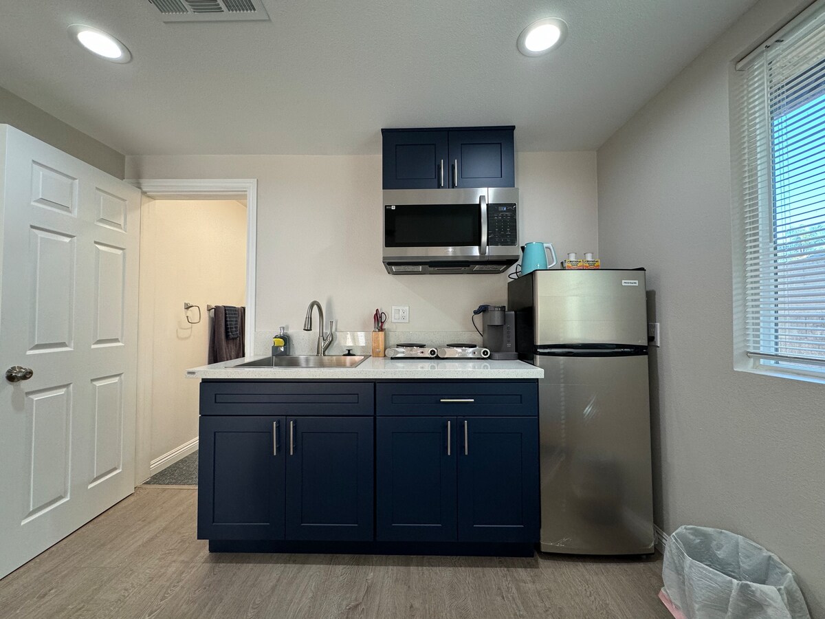 A compact kitchenette is visible, featuring navy blue cabinetry and a light countertop. Appliances include a microwave and a silver refrigerator. Natural light enters through a nearby window, illuminating the space. A doorway leads to a bathroom, partially visible in the background.