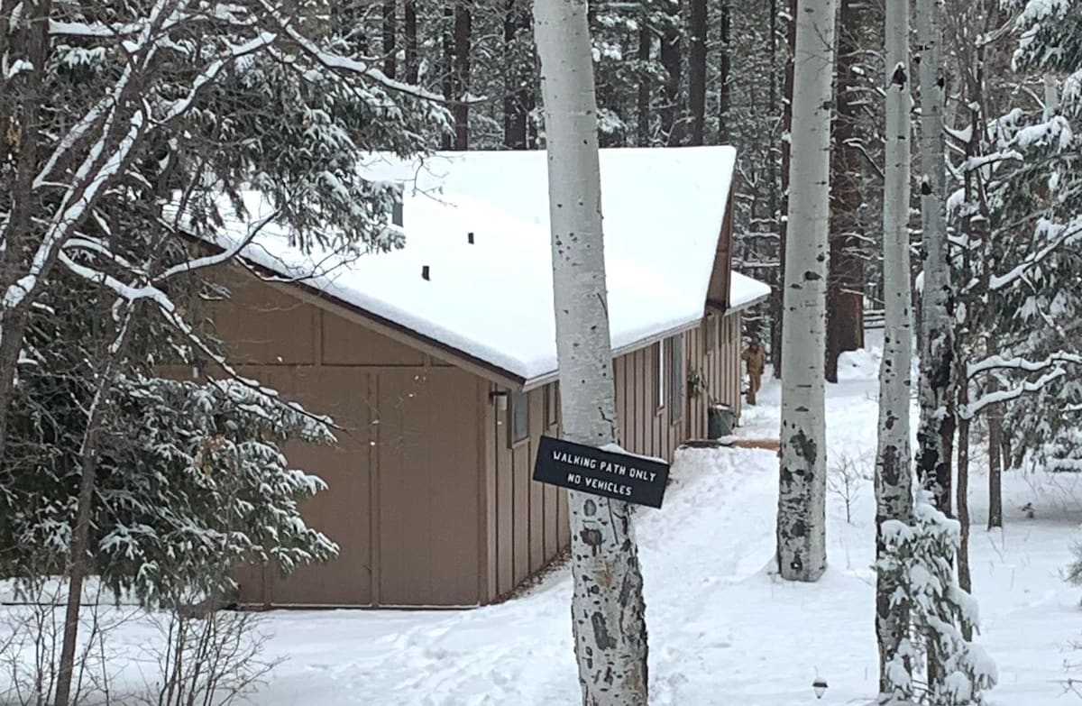 A snow-covered exterior of the Aspen Lodge Bunkhouse is visible, nestled among tall trees. A sign indicating the entrance reads 'Parking Path Only, No Vehicles.' The structure features a sloped roof and is surrounded by a tranquil winter landscape.
