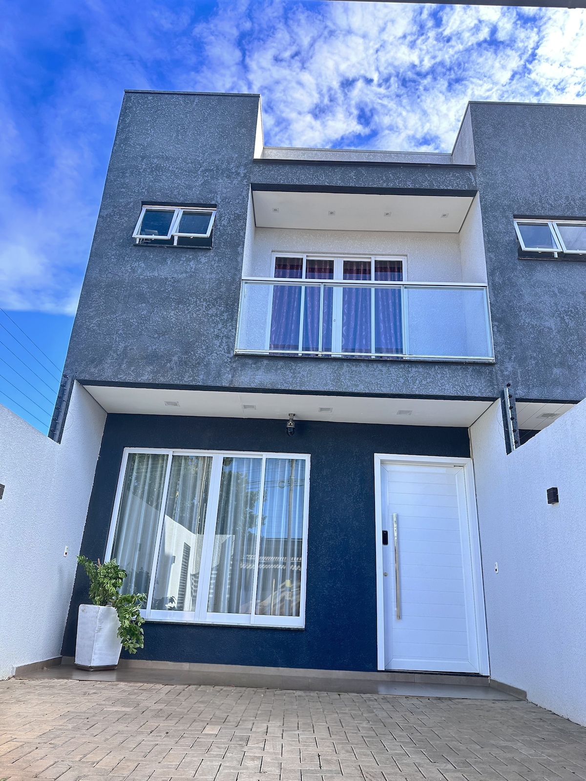 A contemporary two-story house is presented with a sleek façade featuring contrasting dark and light tones. A spacious entryway leads to a modern front door, alongside large windows that allow natural light to enter. A decorative plant sits beside the entrance.