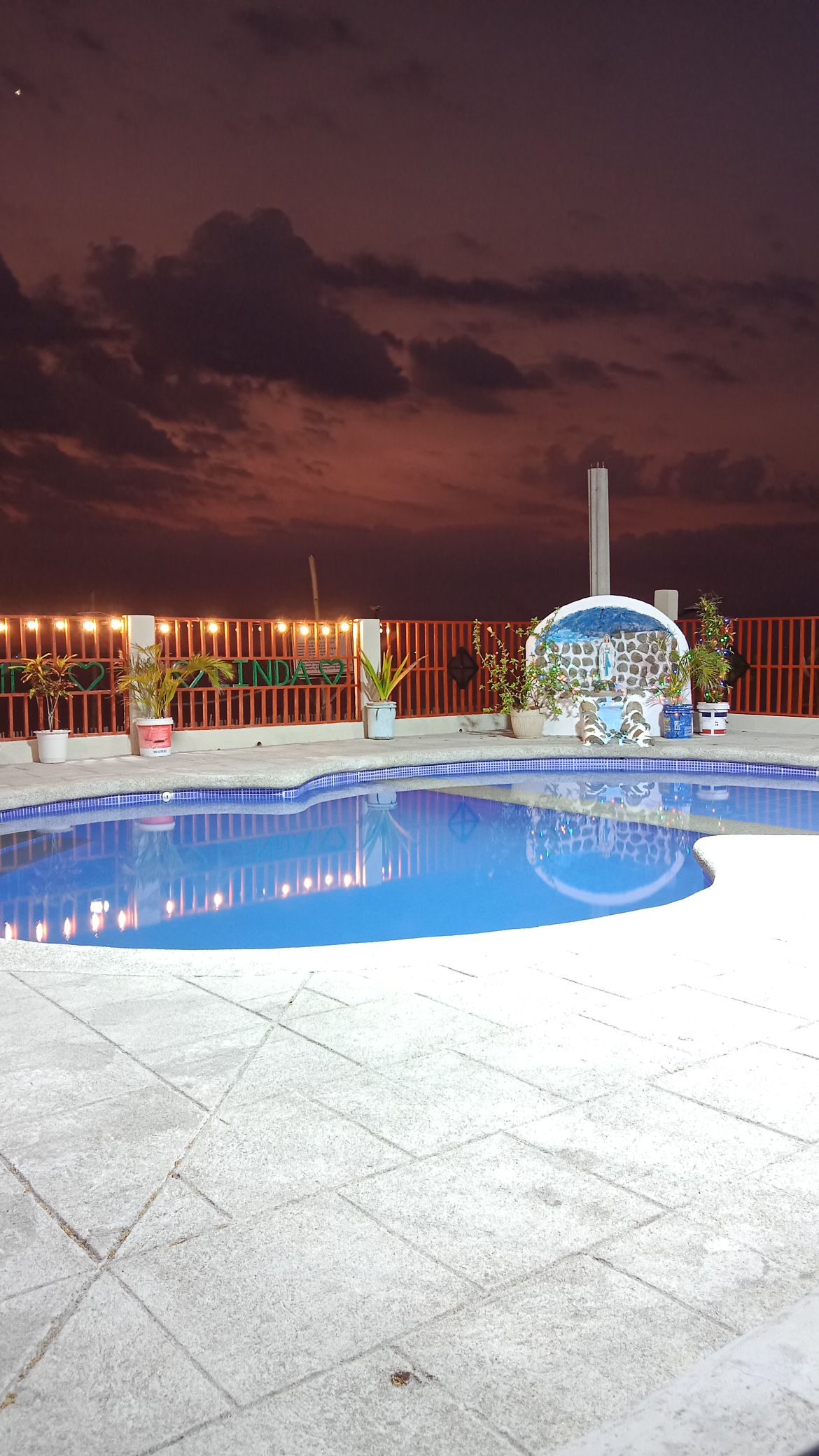 A tranquil pool area is highlighted by clear blue water beneath a dramatic twilight sky. Surrounding planters with greenery add a natural touch, while soft lighting from the fence creates an inviting ambiance.