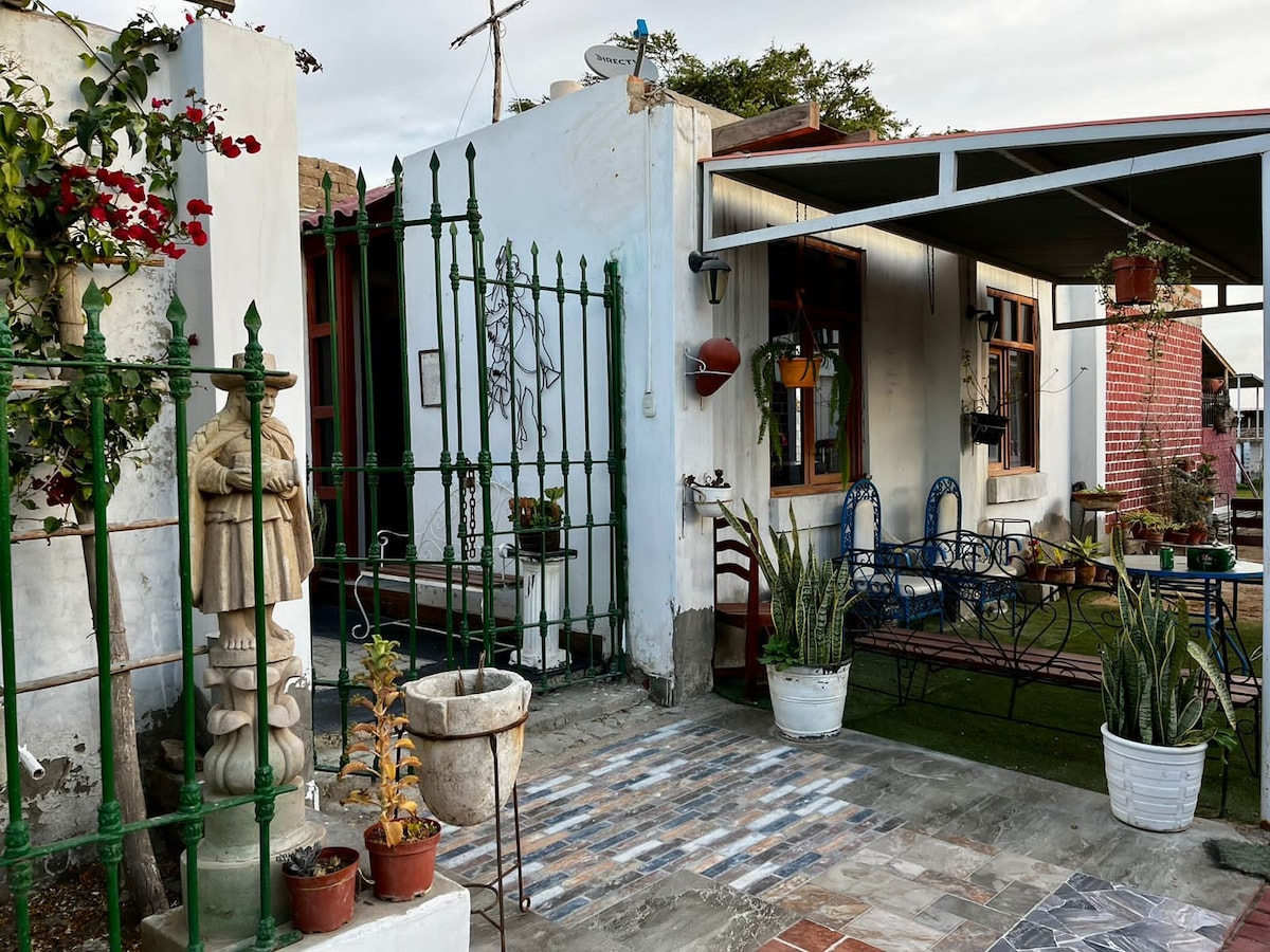 A charming entrance features an ornate green gate leading to a cozy outdoor space. Lush plants and flower pots adorn the area, while a stone statue adds a unique touch. A spacious covered patio is visible, offering seating against a backdrop of a well-kept garden.