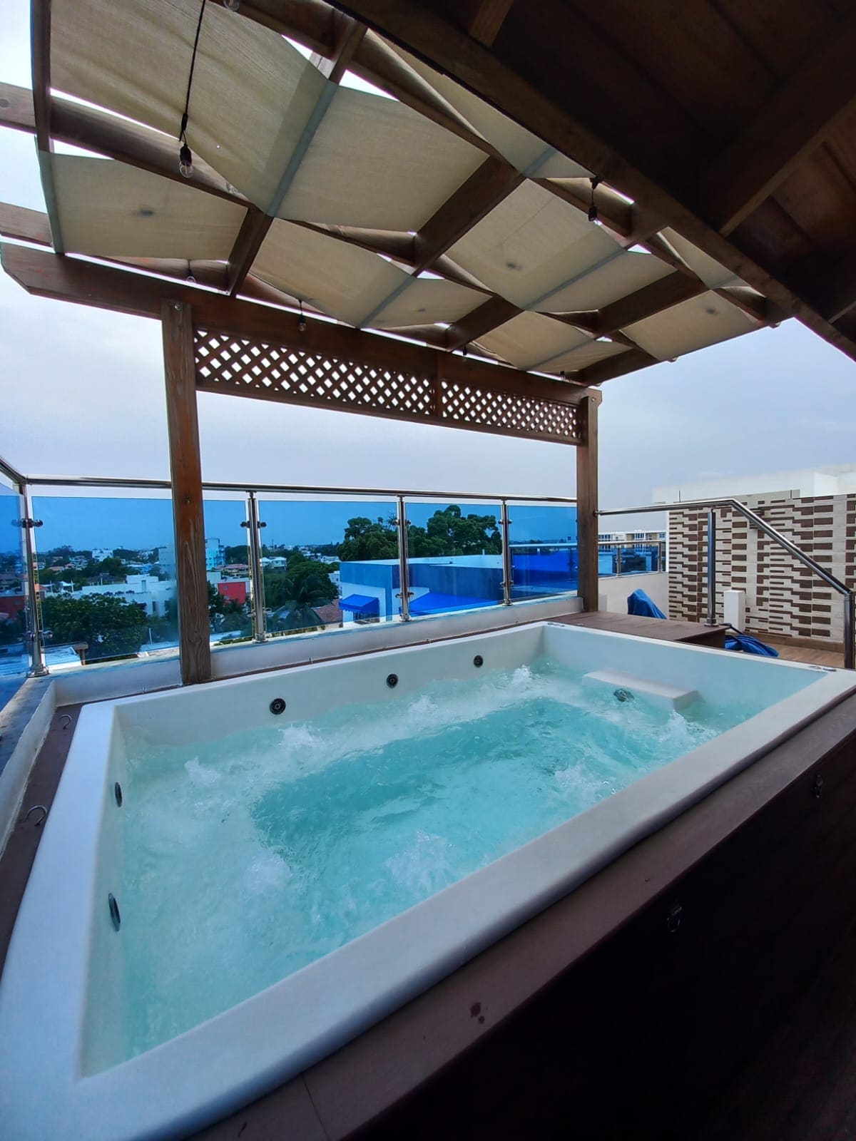 A generous jacuzzi is situated on a private terrace, surrounded by wooden deck and railings. The bubbling water reflects the ambient light, with the city view stretching in the background. A canopy overhead provides shade, enhancing the relaxing setting.