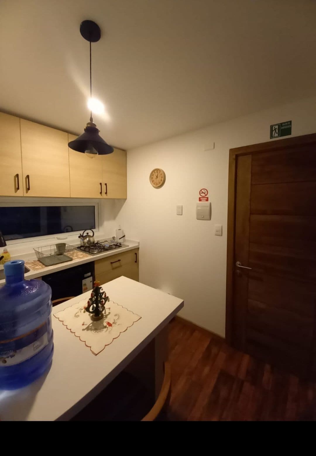 The kitchen features modern wooden cabinetry with a light finish. A sink and a stove are visible under soft lighting. A decorative wall clock adds a touch of style, while a large water dispenser stands on the countertop, enhancing functionality.