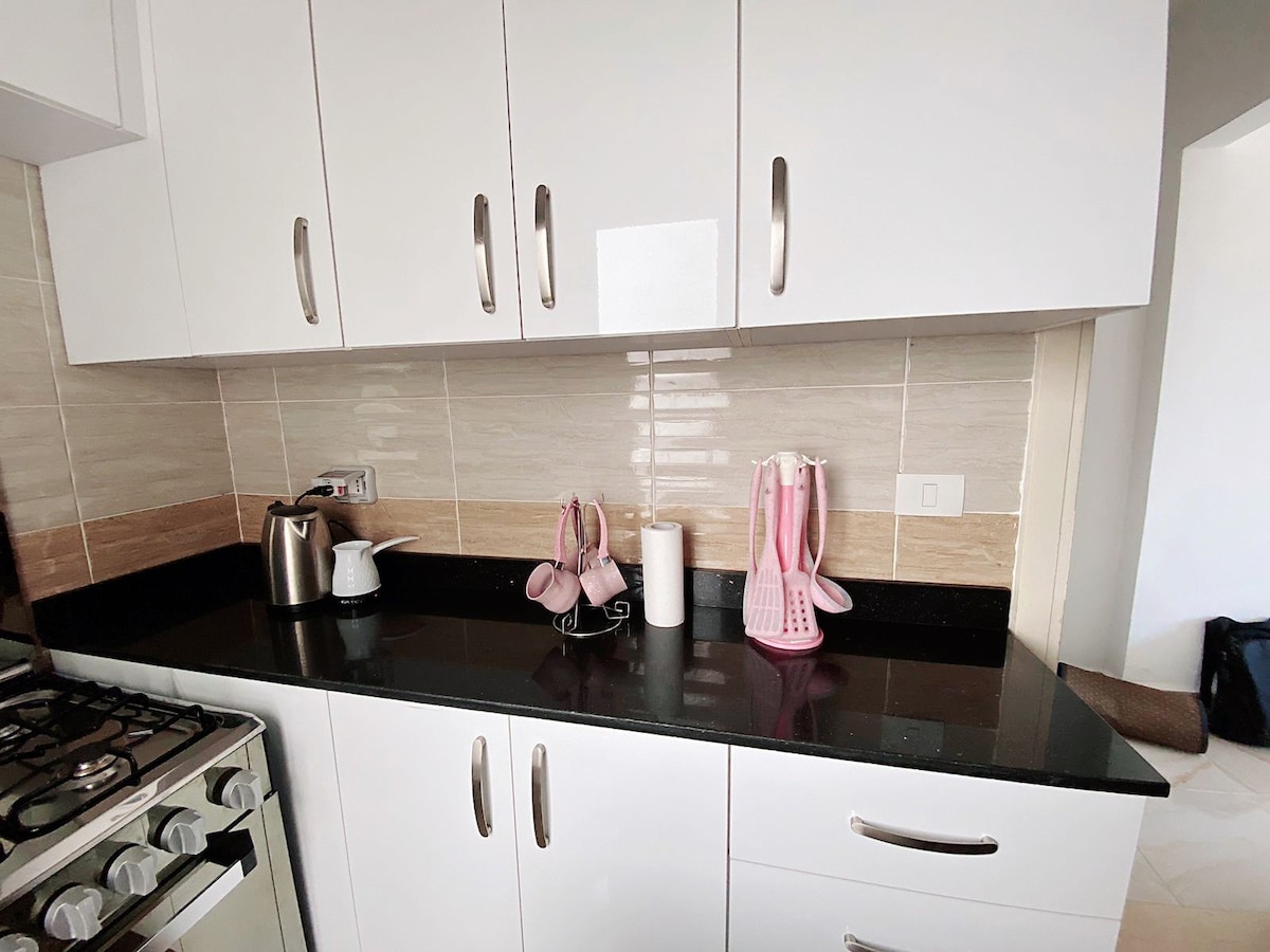 A modern kitchen with white cabinetry and a black countertop is shown. Stainless steel appliances, including a kettle, are placed on the counter, alongside several decorative items in soft pink tones.