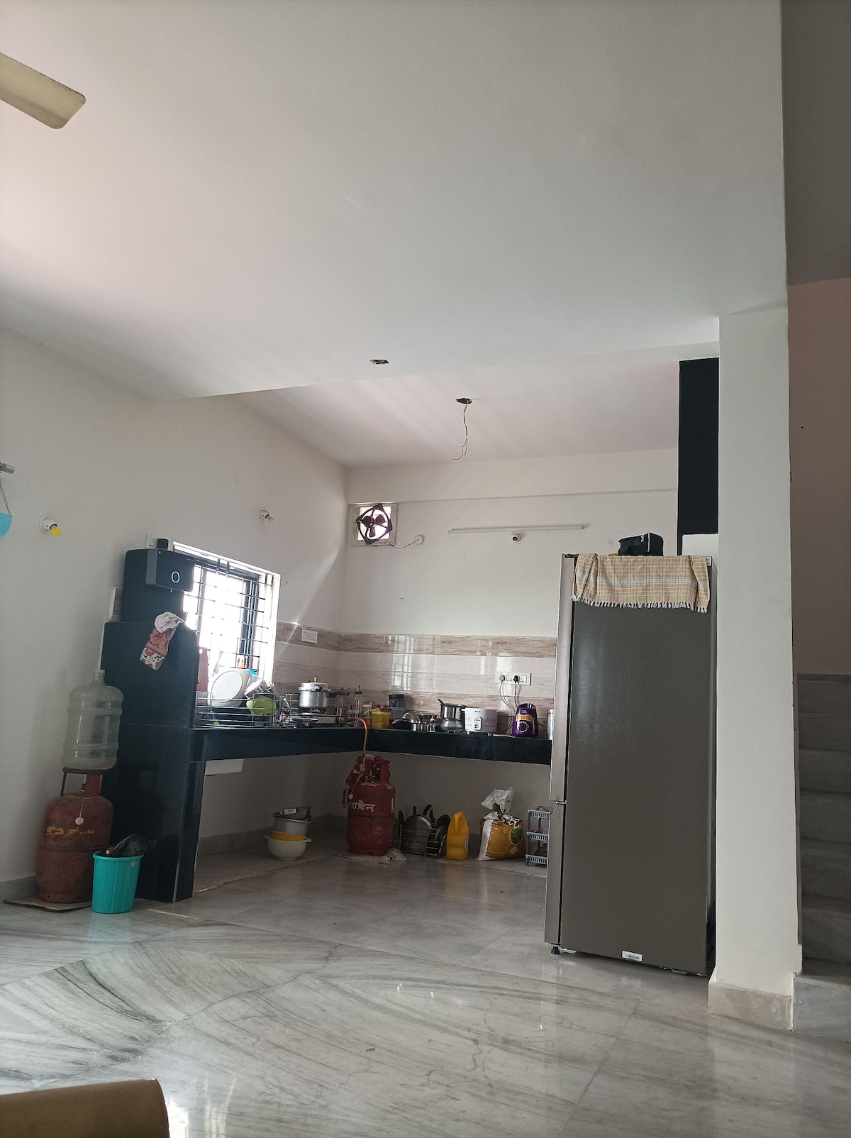 A functional kitchen area features a dark countertop with various kitchen appliances and utensils arranged neatly. A refrigerator stands adjacent to the counter, while light-grey marble flooring enhances the open space. Large windows allow natural light to fill the room.