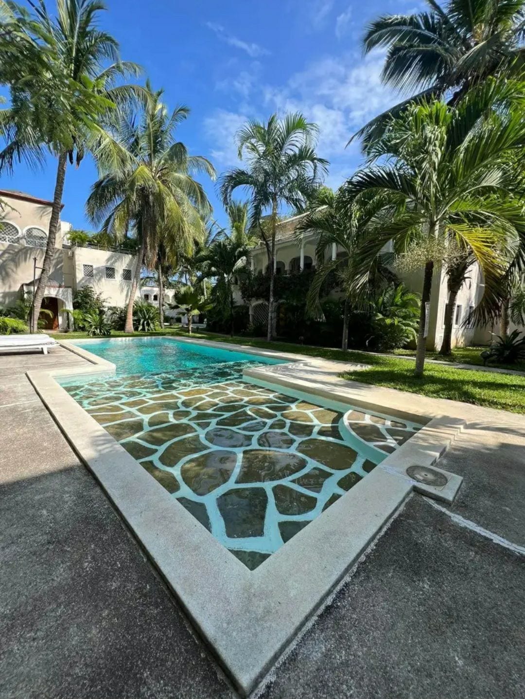 A serene pool is surrounded by lush palm trees and well-maintained gardens. The pool features a unique stone pattern along the edge, contrasting with the clear blue water under a bright sky, creating a refreshing ambiance for relaxation.