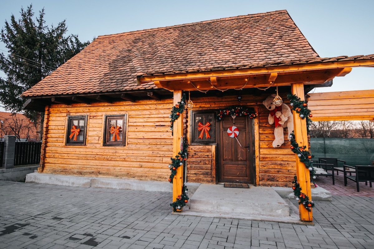 A charming wooden cabin features a traditional design with a sloped roof and wooden accents. Decorative elements, including festive bows and ornaments, adorn the entrance. The porch is spacious, and the surrounding area includes stone pathways and greenery, creating an inviting exterior.