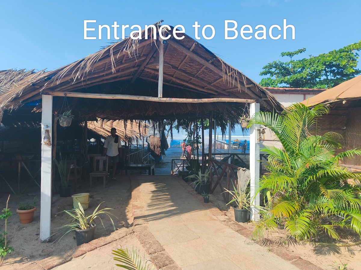 An open entrance leading to the beach is framed by a rustic structure with a thatched roof. Pathways lined with potted plants guide the way toward the sandy shore, visible in the background under a clear blue sky.