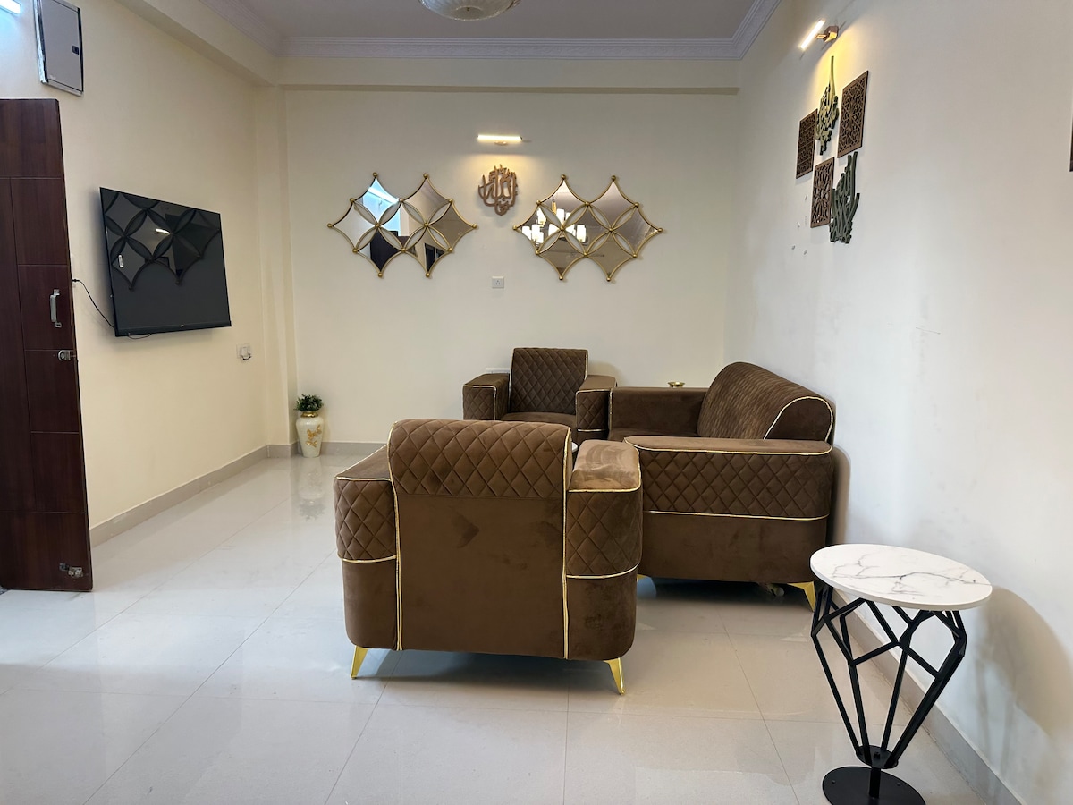 A cozy living area features a pair of stylish, brown upholstered sofas arranged around a small marble-top side table. Elegant wall mirrors reflect light and add depth to the space. A television mounted on the wall complements the modern decor.