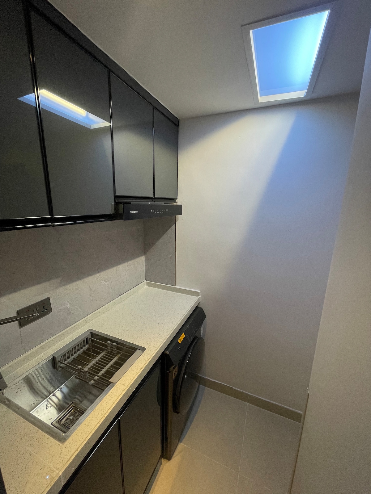 A compact kitchenette is presented with sleek black cabinetry and modern fixtures. A small stainless steel sink is situated next to an induction cooktop. An in-unit washer and dryer are positioned nearby, under a ceiling light that illuminates the space with a soft glow.