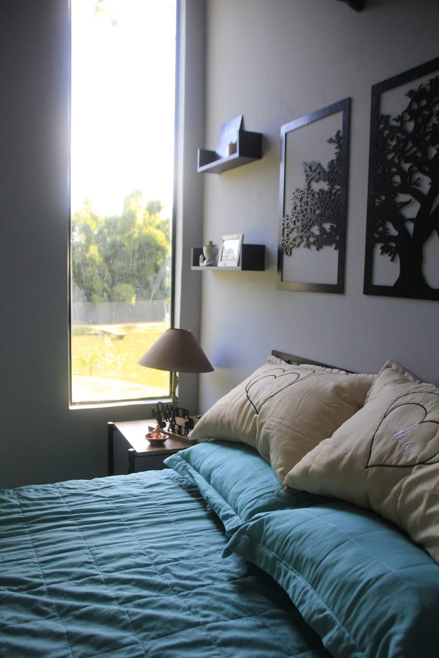 A cozy bedroom is presented, featuring a spacious bed with light blue bedding and decorative pillows. A bedside table holds a lamp, while framed artwork adorns the wall. Natural light filters through a large window, illuminating the serene outdoor view.