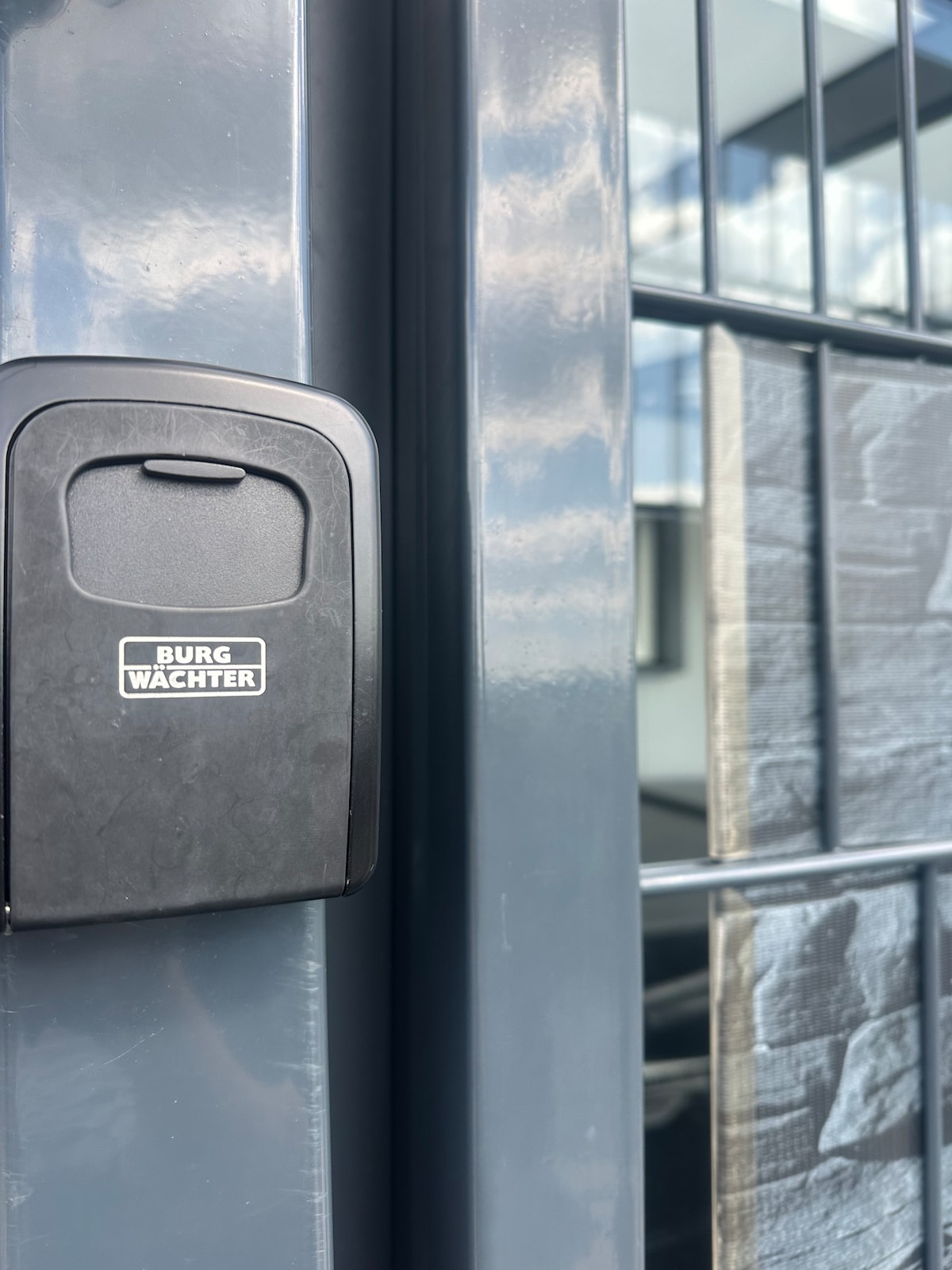 A close-up view of a secure key box is presented, featuring a sleek black design and the brand name 'BURG WACHTER.' The box is mounted on a dark exterior wall, with glass panels visible in the background, indicating an entrance to the property.