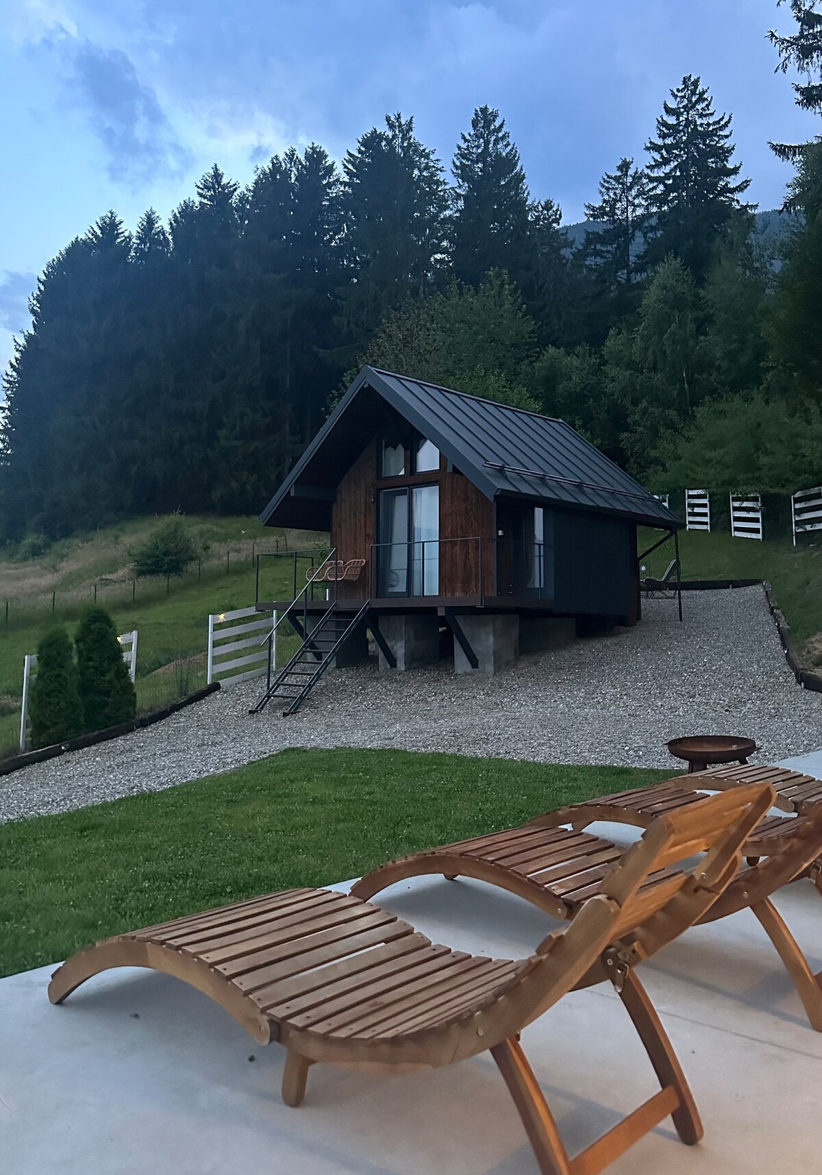 A charming cabin is set against a backdrop of tall trees, featuring a sloped roof and wooden accents. The gravel path leads to the entrance, and two wooden loungers are positioned on the patio area, inviting relaxation amidst the natural surroundings.